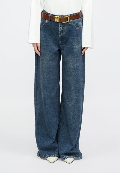 Luźne, szerokie spodnie dżinsowe w kolorze niebieskim o wyblakłej teksturze, zestawione z brązowym skórzanym paskiem z pozłacaną klamrą oraz białą, długorękawową bluzką.