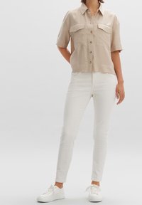 Beige kortärmad skjorta med knappar och två bröstfickor, matchad med slim-fit vita jeans och vita sneakers. Slät tygtextur.