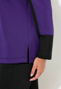 Lila topp med svarta ärmar, med en sidodelning och en mjuk tygkvalitet. Synlig sömdetalj vid fåll och ärmkanter.