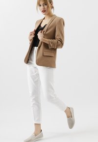 Lichtbruine blazer over een zwarte kanten top, gecombineerd met witte slanke broeken en geperforeerde lichtgrijze instapper schoenen. Gladde stoftextuur.