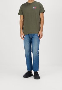 Olivově zelené bavlněné tričko s malým barevným logem na levé straně hrudi, v kombinaci se středně modrými džínami a černými teniskami.