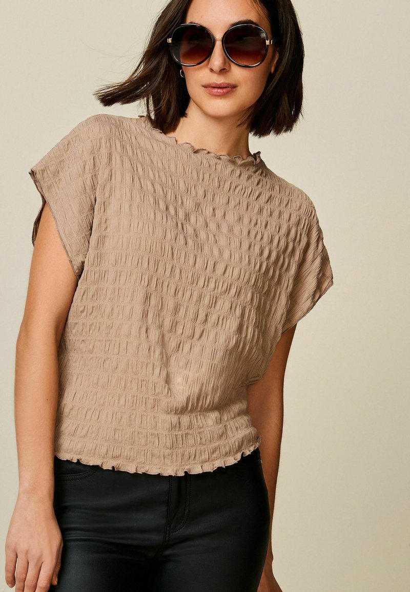 Beige texturierte Bluse mit kurzen Ärmeln, gerüschtem Saum und lockerem Schnitt, kombiniert mit schwarzen figurbetonten Hosen und übergroßen Sonnenbrillen.