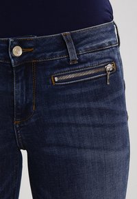 Jeans de mezclilla azul oscuro con un bolsillo con cremallera en la parte frontal. La textura de la tela es suave y ligeramente desgastada, con detalles de costura y herrajes visibles.