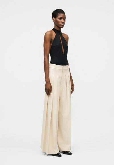 Femme portant un haut noir sans manches à col licou avec une découpe à l'avant, un pantalon beige taille haute à jambes larges, et des chaussures noires pointues.
