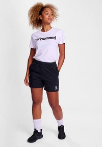 Biała koszulka z czarnym logo "hummel", zestawiona z czarnymi szortami. Model nosi białe skarpetki i czarne buty sportowe. Tkanina o fakturze.