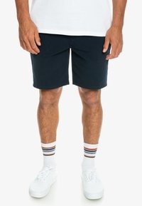 Shorts bleu marine en mélange de coton, avec une coupe décontractée et des poches latérales. Associés à des baskets blanches et des chaussettes montantes rayées.