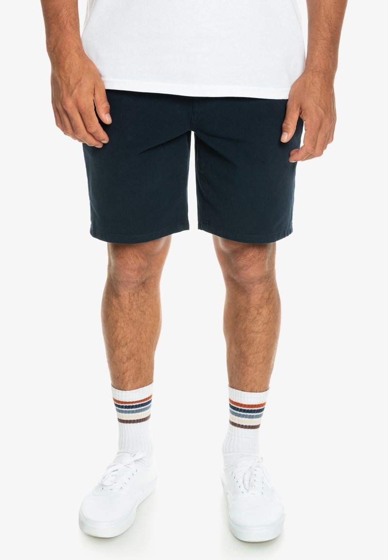 Shorts bleu marine en mélange de coton, avec une coupe décontractée et des poches latérales. Associés à des baskets blanches et des chaussettes montantes rayées.