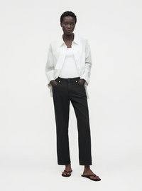 Mujer de pie con las manos en los bolsillos, lleva camisa blanca abotonada sobre camiseta blanca, pantalones negros y sandalias negras de chancla.