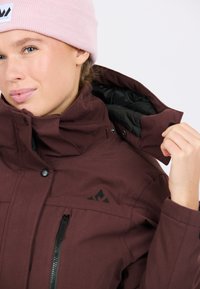 Bordeaux geïsoleerde jas met zwarte voering, ritszak en hoge kraag. Gecombineerd met een lichtroze ribgebreide beanie.