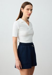 Witte geribde polo met een golvende kraag, gecombineerd met marineblauwe shorts met trekkoord, voorzien van voorzakken en een gladde textuur.