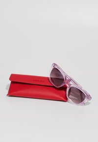 Gafas de sol transparentes rosas con lentes degradados junto a un estuche de cuero rojo grabado con "GUESS". Texturas suaves y diseño angular.