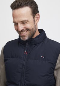 Mørkeblå polstret vest med høy krage og glidelås foran; har et lite logo med røde, hvite og blå detaljer på brystet.