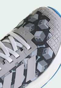 Scarpa sportiva grigia con tomaia a motivo esagonale, dettagli grigi testurizzati, lacci bianchi e una robusta suola bianca con dettagli blu.