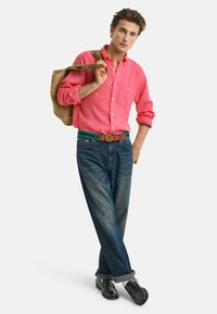 Camicia di lino rossa, jeans blu leggermente sbiaditi, cintura di pelle marrone e scarpe nere. Tiene una borsa color sabbia su una spalla.