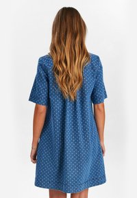 Robe en denim bleu avec des manches courtes, une coupe décontractée et un motif à petits pois blancs. L'ourlet est légèrement arrondi au bas.