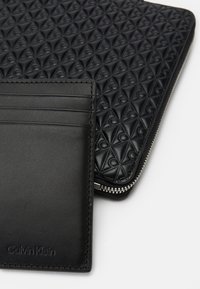 Porte-cartes en cuir noir avec quatre fentes à l'avant, associé à une pochette zippée noire texturée présentant un motif géométrique.