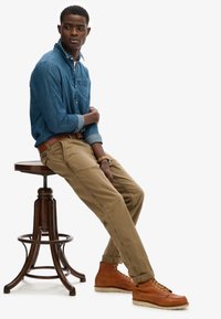 Denim skjorte med knapper, beige chinos og brune snørebåndsstøvler. Model sidder på en træbarstol og viser en afslappet pasform og en casual stil.