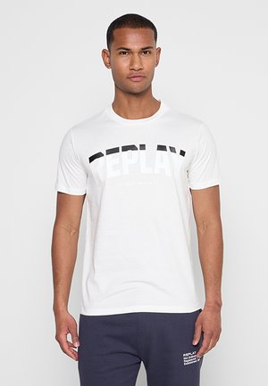 Mann, der frontal steht und ein weißes T-Shirt mit dem Schriftzug "REPLAY" sowie eine dunkle Hose mit weißem Text am oberen Oberschenkel trägt.