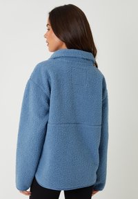 Fleece pullover i lys blå, med krave, lange ærmer og en tekstureret overflade. Bagudseende viser stikningsdetaljer og løs pasform.