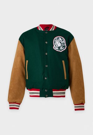 Un homme porte une veste universitaire verte à manches marron, un polo rayé, un short beige, des chaussettes blanches, des baskets et une casquette rose.