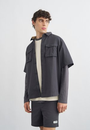 Noor mees, kandes kihilist tumerohelist särki, beeži kampsunit ja tumerohelisi lühikesi pükse, seisab valge ühevärvilise tausta ees ja vaatab küljele.