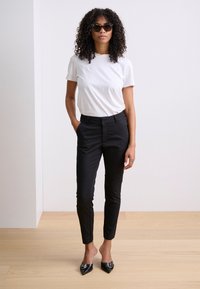 Pantalons noirs ajustés, t-shirt blanc à col rond et escarpins noirs à bout pointu. Le mannequin porte des lunettes de soleil oversized noires et a les cheveux bouclés.