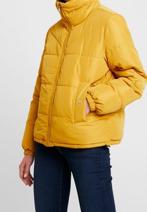 Personne portant une doudoune matelassée jaune avec un col montant et des poches latérales, associée à un jean bleu foncé.