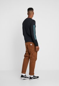 Hombre de pie de espaldas, vestido con una camiseta de manga larga negra con texto azul en la manga, pantalones marrones, calcetines blancos y zapatillas negras.