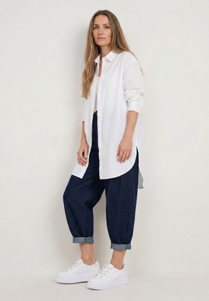 Frau steht an der Wand, trägt ein langes weißes Hemd mit Knöpfen, hochgekrempelte dunkelblaue Hosen und weiße Sneaker.