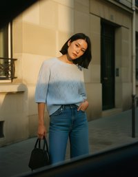 Gaiši zils adīts džemperis ar rakstainu struktūru, apvienots ar augsta vidukļa zilajiem džinsiem. Modelis tur nelielu melnu rokassomu. Fona attēlā redzama akmens siena.