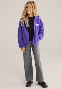 Violeta flīsa jaka ar kapuci, ar rāvējslēdzēja aizdari un teksturētu audumu. Apvienota ar melnu krekli un pelēkiem platiem džinsiem. Melnas kurpes.