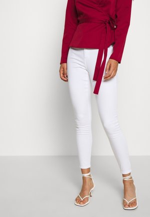 Kvinde iført hvide skinny jeans, hvide rem-sandaler med hæl og en langærmet rød slå-om top med bindebånd i taljen.