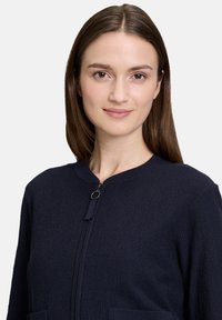 Marineblaue Zip-Jacke aus strukturiertem Stoff, mit rundem Halsausschnitt und zwei Fronttaschen. Verfügt über einen zentralen Reißverschluss mit einem Ziehgriff.