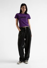 Purpurowy skrócony t-shirt z czarnym graficznym tekstem, w połączeniu z luźnymi czarnymi spodniami z sztucznej skóry i czarnymi trampkami z białą podeszwą.