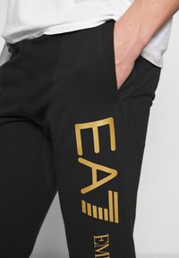 Svarta sweatpants med guldtryck, med en sidoficka och en mjuk textur. Logotypen på benet säger "EA7" och "EMEA."