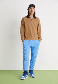 Bruine polo sweatshirt met lange mouwen en zwarte kraag, gecombineerd met lichtblauwe sweatpants en blauw-witte sneakers. Eenvoudige, casual outfit.