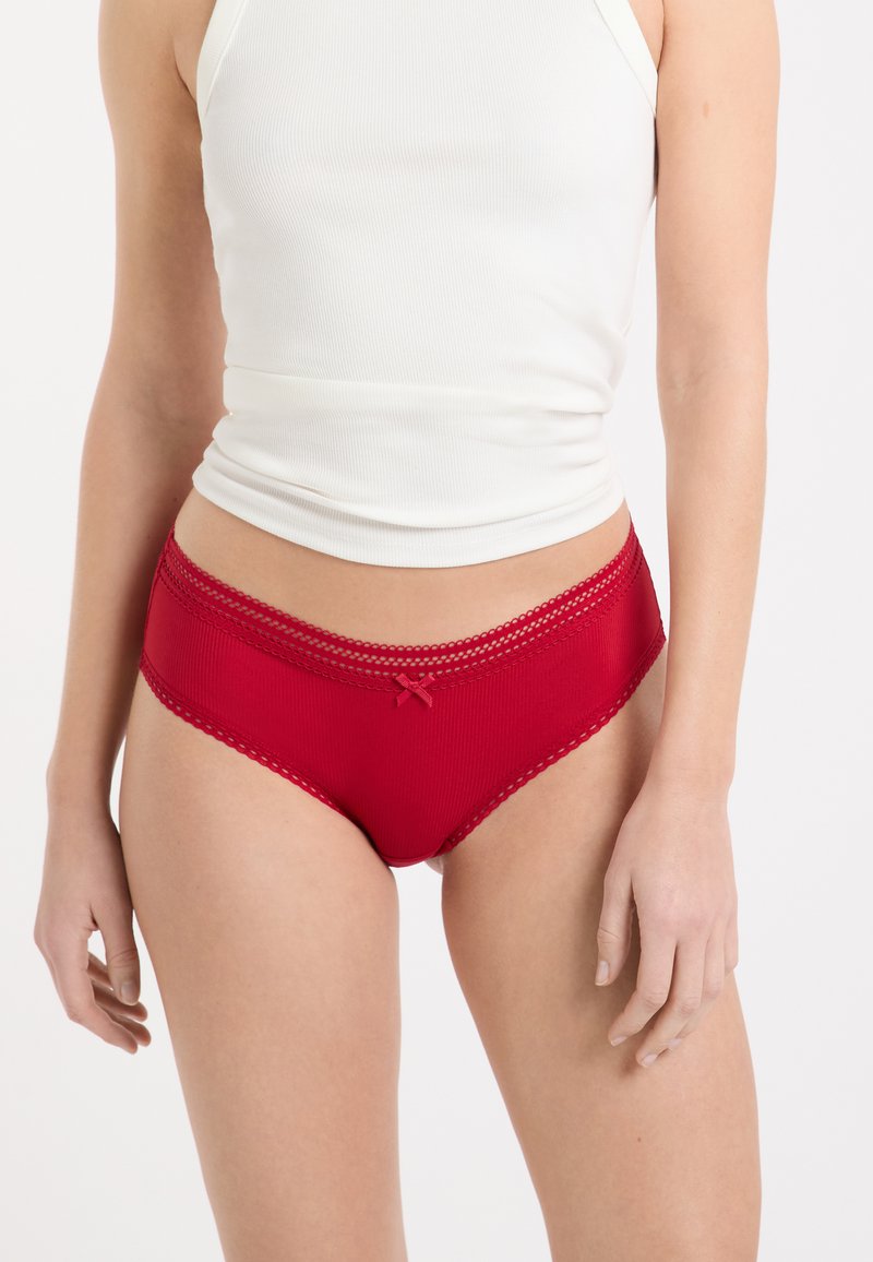 Culotte en coton rouge avec une texture côtelée, bordée de dentelle et ornée d'un petit nœud à l'avant. Conception taille haute, bords lisses.