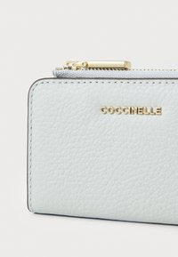 Portefeuille en cuir bleu clair avec fermeture éclair et quincaillerie en métal doré. Présente un logo embossé "Coccinelle" et une surface texturée visible.
