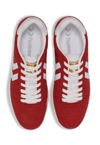 Red suede sneakers with white accents, featuring a lace-up design, perforated vamp for breathability, and a gold logo detail.