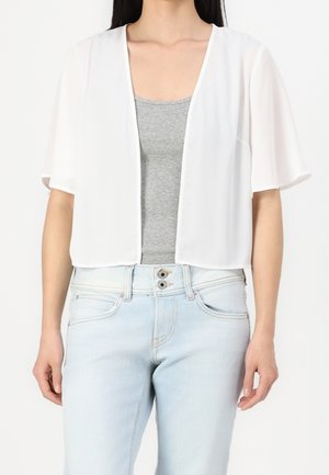 Frau trägt eine transparente weiße, vorne offene Kurzarm-Strickjacke über einem grauen Tanktop und hellblauen, hoch taillierten Jeans.