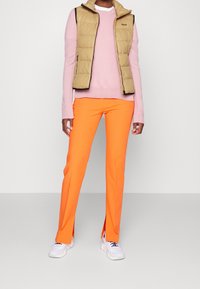 Béžová zateplená vesta přes světle růžový svetr, doplněná jasně oranžovými kalhotami a bílými teniskami, s modrými akcenty a bočními rozparky.