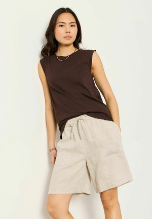 Kobieta w ciemnobrązowej bluzce bez rękawów i beżowych szortach ze ściągaczem, stojąca na tle jednolitej białej ściany.
