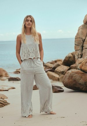 Kobieta o długich blond włosach stoi boso na piaszczystej plaży w pobliżu skał, ubrana w pasiasty top bez rękawów i szerokie spodnie z koronkowymi detalami.