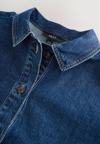 Cămașă din denim cu guler ascuțit, cusături contrastante și două nasturi închiși la culoare vizibili în apropierea gulerului și a zonei pieptului.