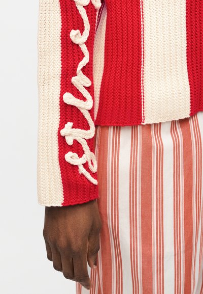Σκούρο δέρμα χέρι δίπλα σε κόκκινο και κρεμ πλεκτό μανίκι πουλόβερ με ανάγλυφο κείμενο με καλλιγραφική γραφή, συνδυασμένο με κόκκινη και λευκή κάθετα ριγωτή φούστα.
