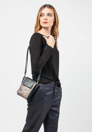 Kleine Umhängetasche in Schwarz und Beige, mit einer oberen Klappe, goldenen Beschlägen und einer bedruckten Textur mit Schriftzügen. Getragen zu einem schwarzen Langarmoberteil.