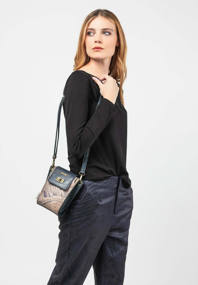 Piccola borsa a tracolla nera e beige, con un flap superiore, hardware dorato e una texture stampata con dettagli in scrittura. Indossata con un top a maniche lunghe nero.