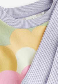 Värikäs palloneule, jossa on liila ribbikaulus. Ominaisuuksiin kuuluu pehmeä, teksturoidu kangas ja päällekkäisten muotojen kuvio pinkissä, oranssissa ja vihreässä.