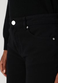 Jeans in denim nero con vestibilità slim, dotati di accessori in metallo argentato, una tasca frontale e tessuto testurizzato. Le cuciture sono visibili in toni contrastanti.