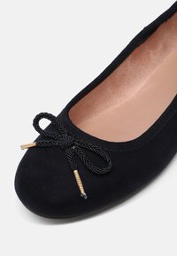 Ballerine noire avec nœud tressé et bouts dorés, avec un bout arrondi et une doublure intérieure beige.